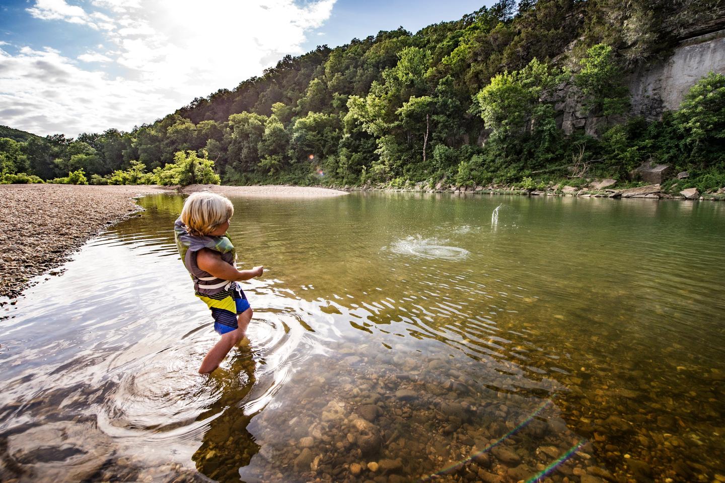 Child splashing in a river with a life vest under a cloudy sky.
