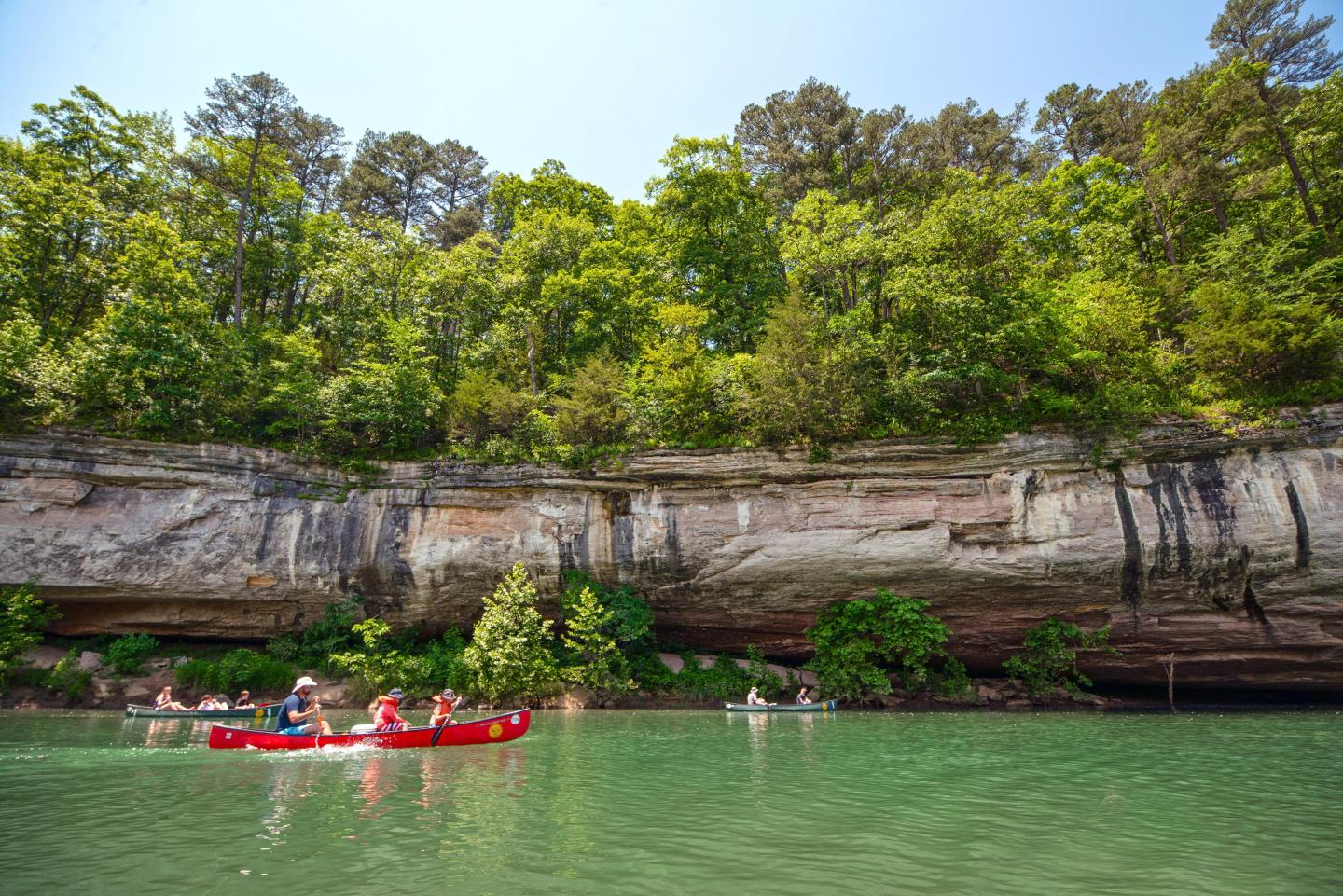 Canoeists paddle on a river near a rocky, tree-lined cliff under a clear blue sky.