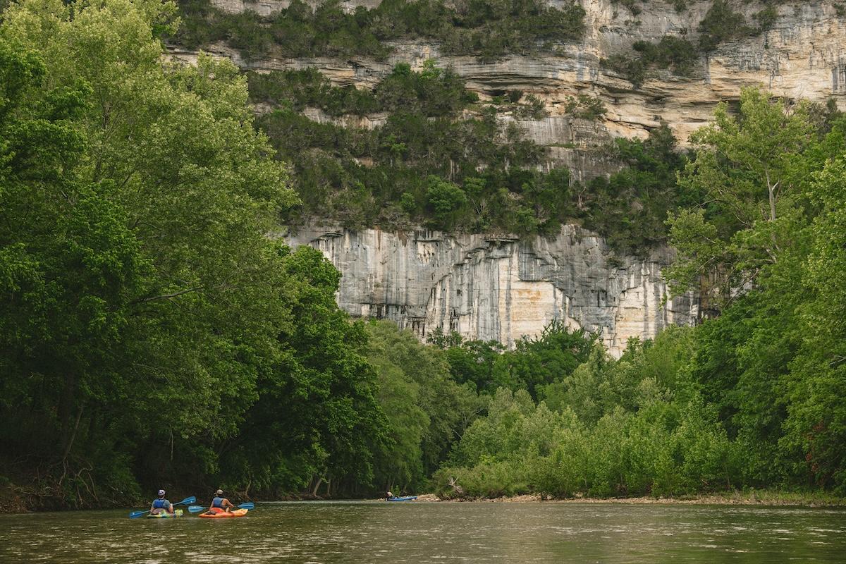 Kayakers paddle along a river surrounded by lush green trees and rocky cliffs.