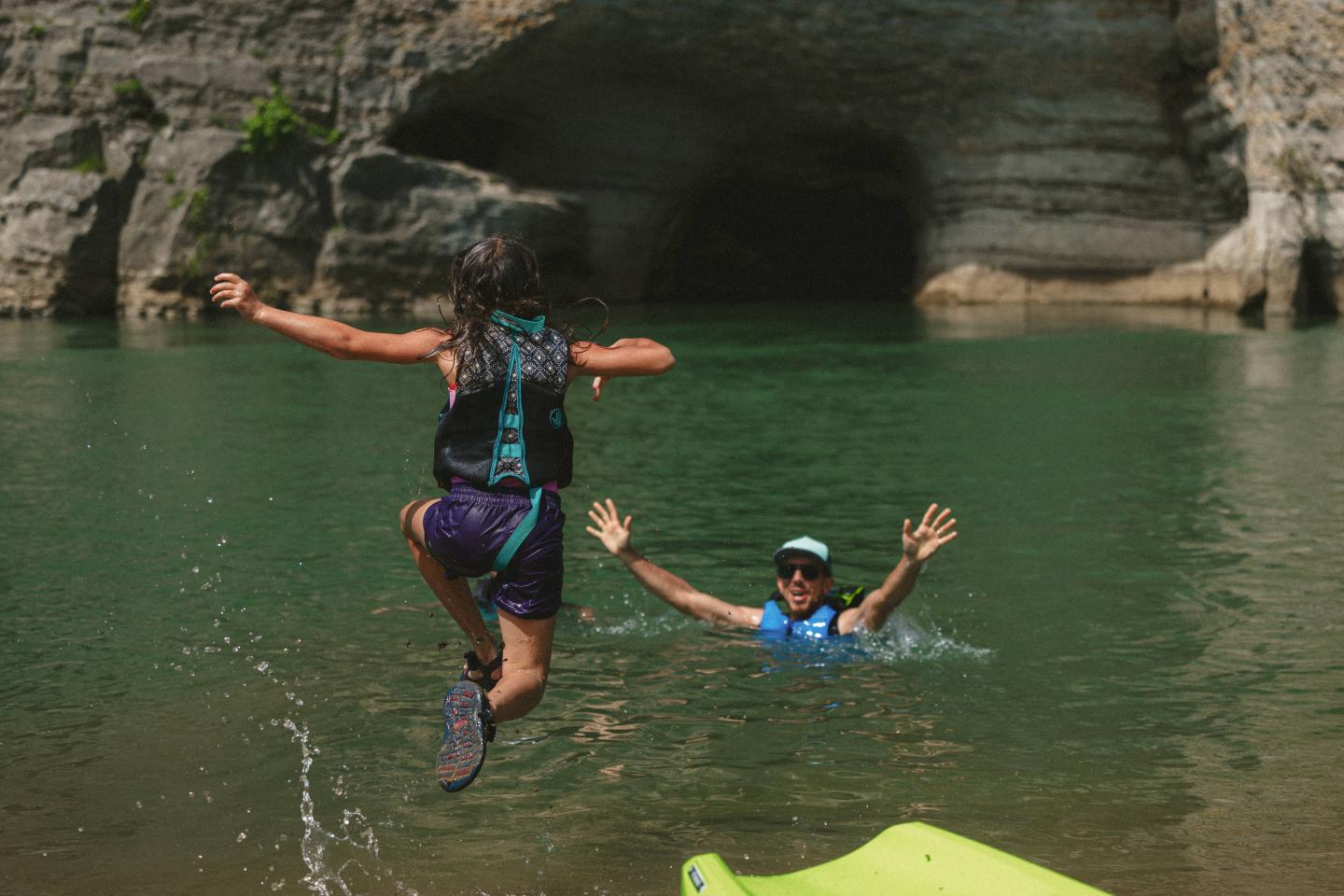 Child jumps into water towards an adult in a rocky cove, both smiling.