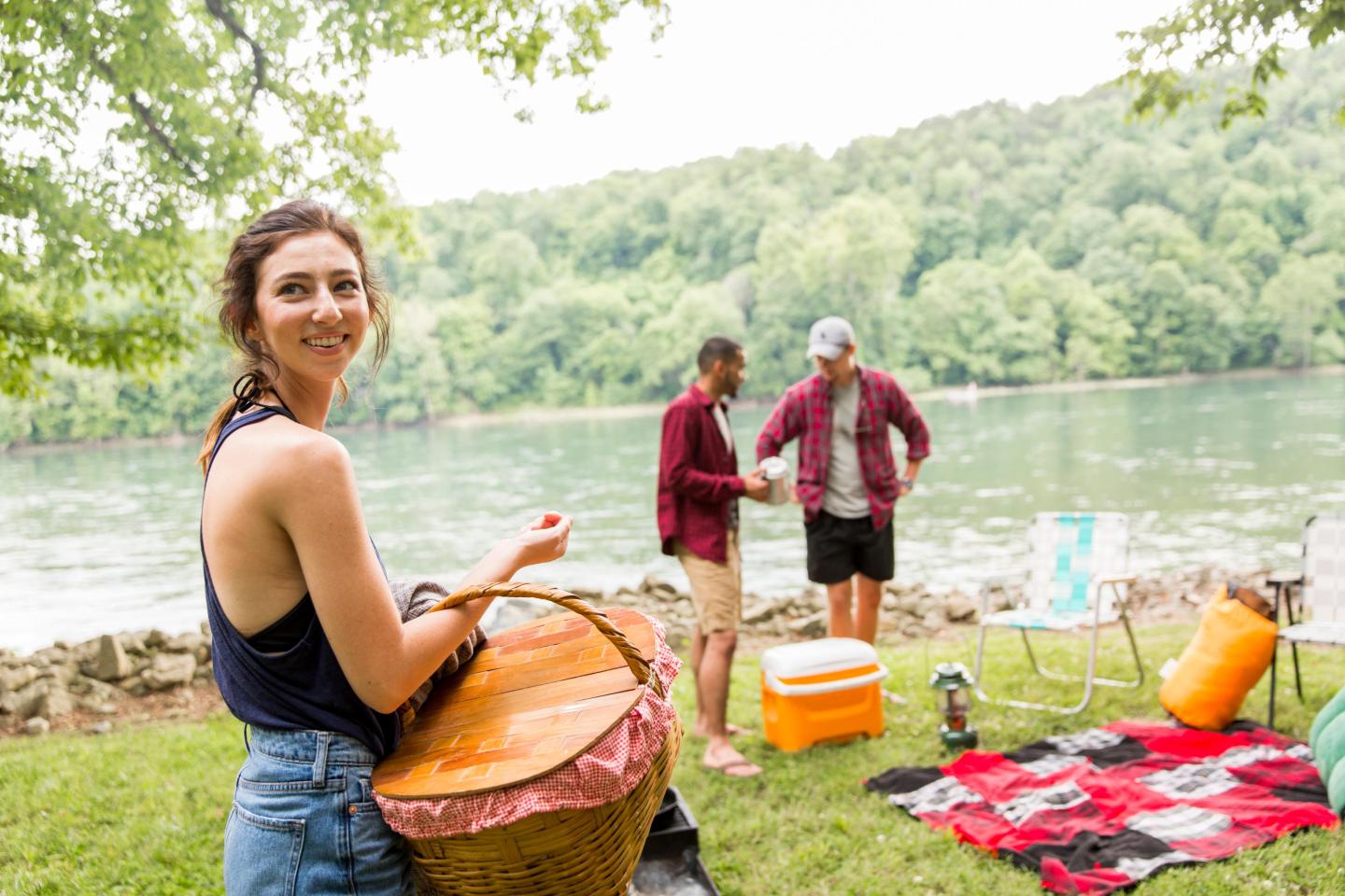 A woman holding a picnic basket by a river, two people in the background, lush trees surrounding.