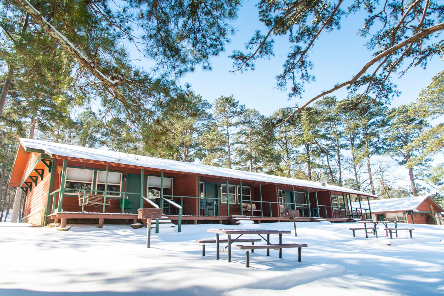 Snow-covered lodge with picnic tables and pine trees on a sunny day.