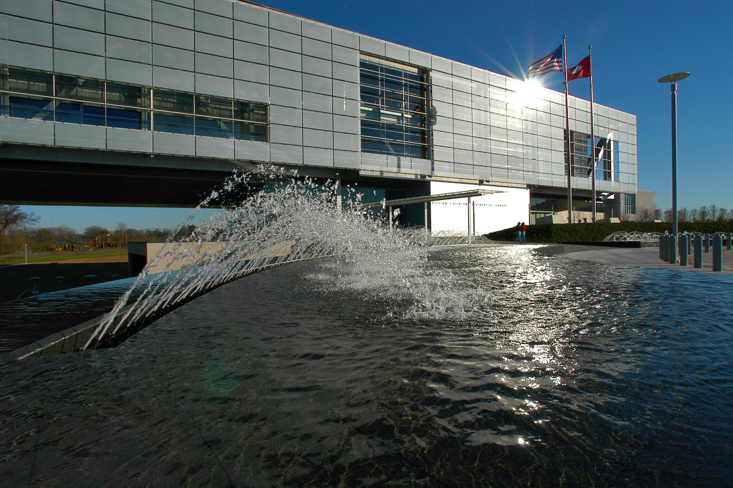 Modern building with a reflective pool and fountain, flags above, under a clear blue sky.