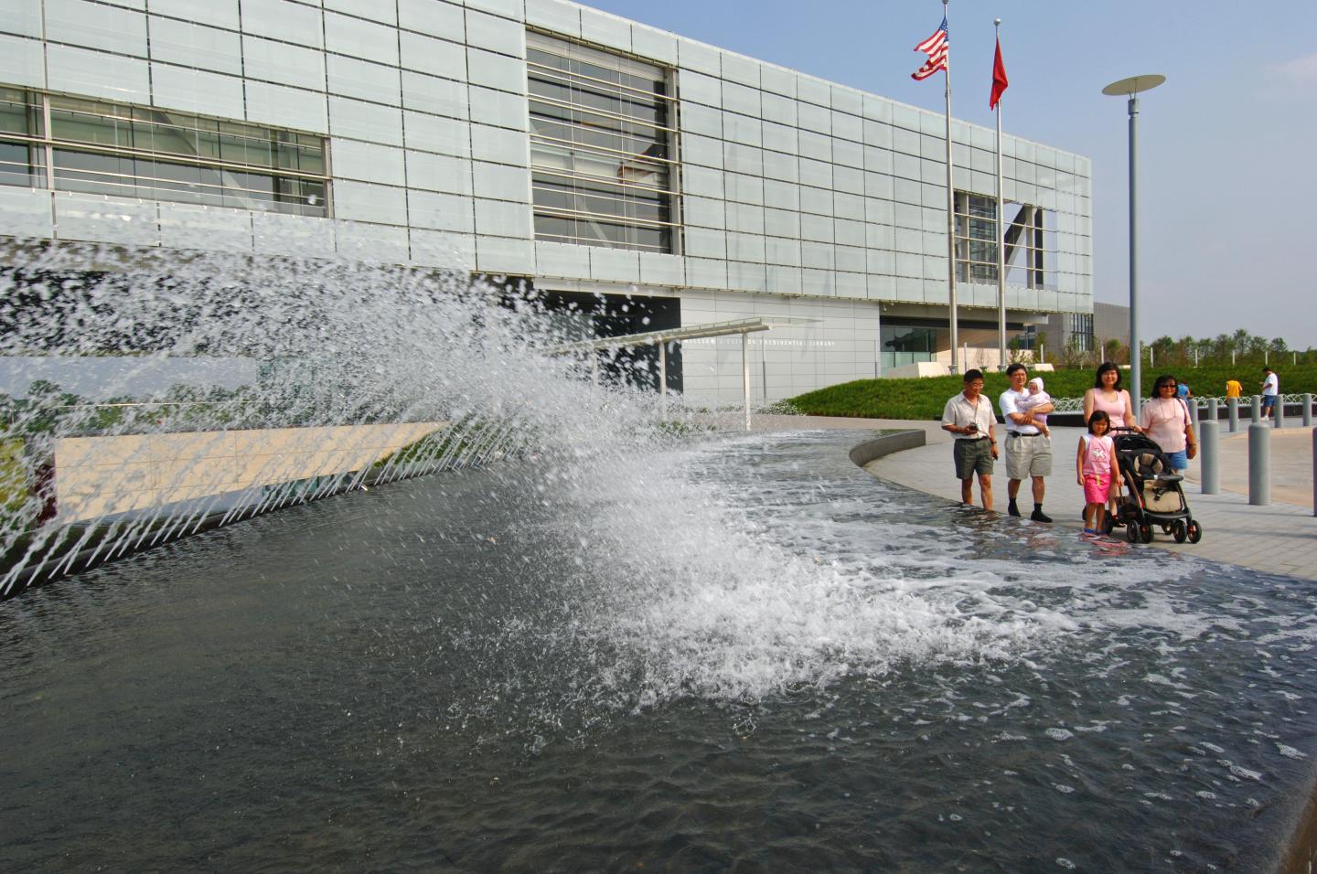 Water fountain spraying near a modern building with flags, people watching nearby.