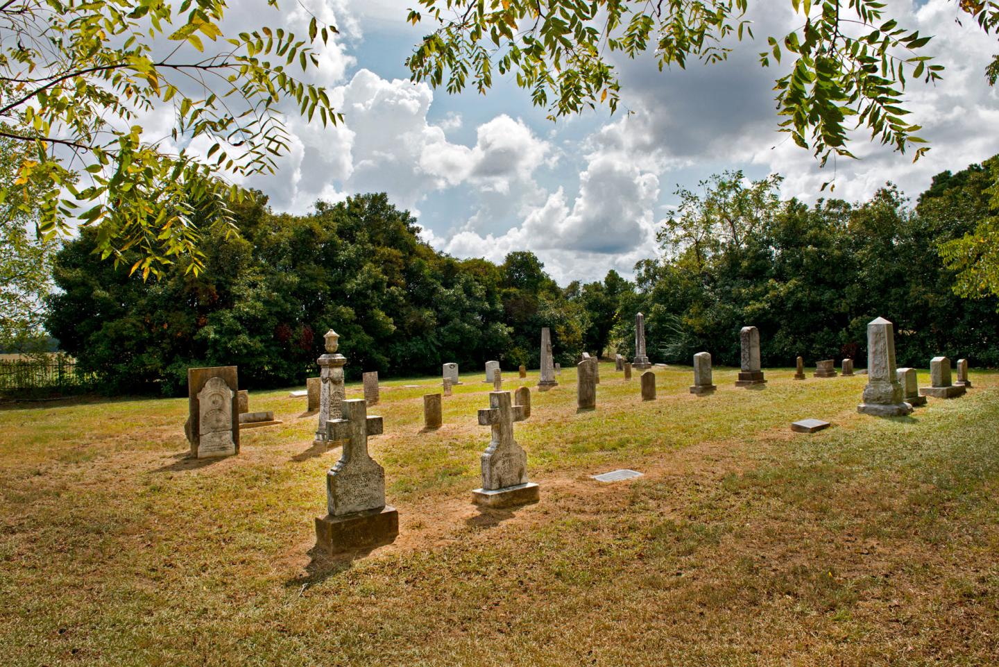 Grassy cemetery with old tombstones, trees, and clouds in the background.