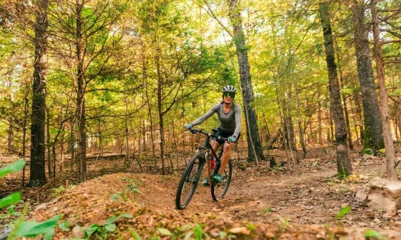 Cyclist rides through a sunny, wooded trail surrounded by green and brown foliage.