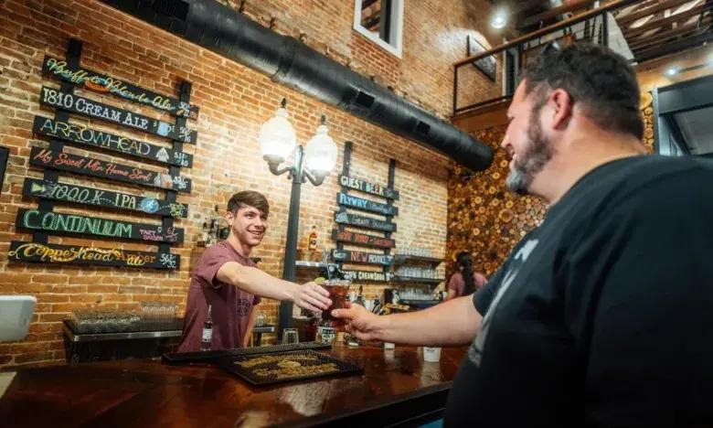 Bartender handing a drink to a smiling customer in a lively, rustic bar.