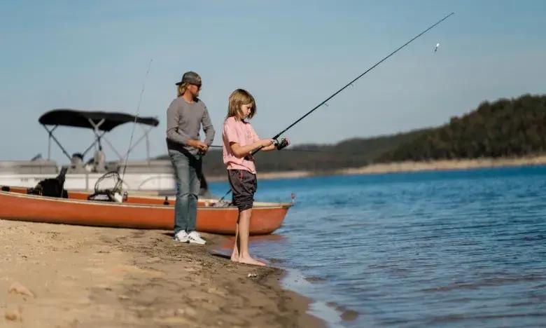 Father and child fishing by a lake, boat nearby, sunny day.