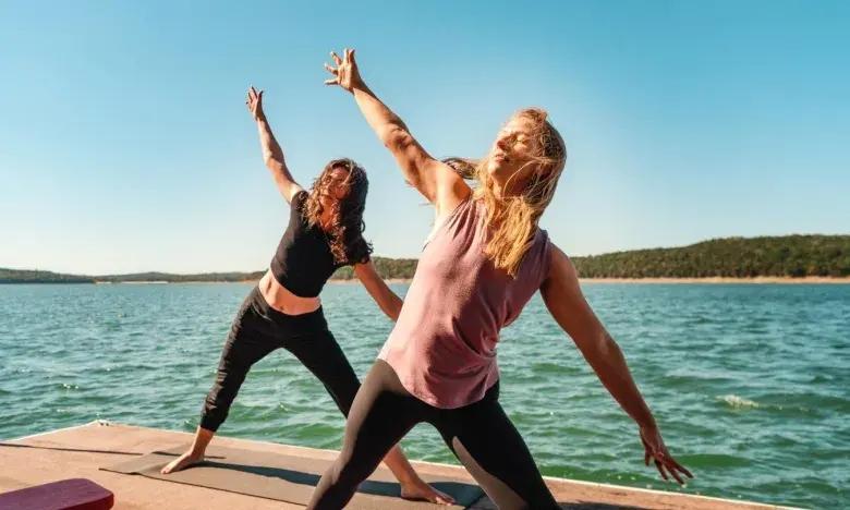 Two people doing yoga on a lakeside deck under a clear blue sky.