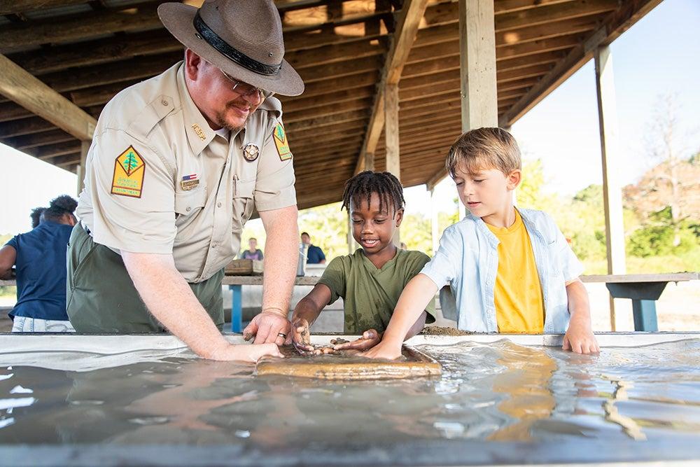 Park ranger and two kids touch a turtle under a covered outdoor area.