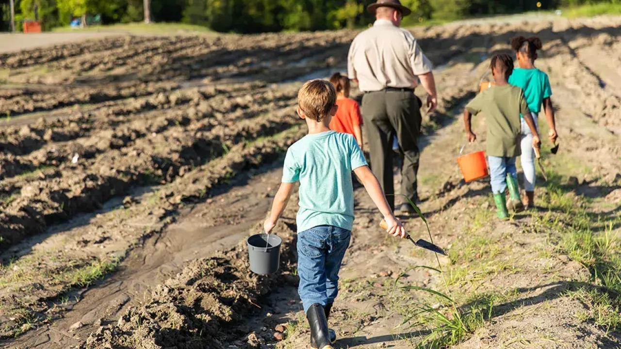 Children walking in a field with buckets and tools, accompanied by an adult.