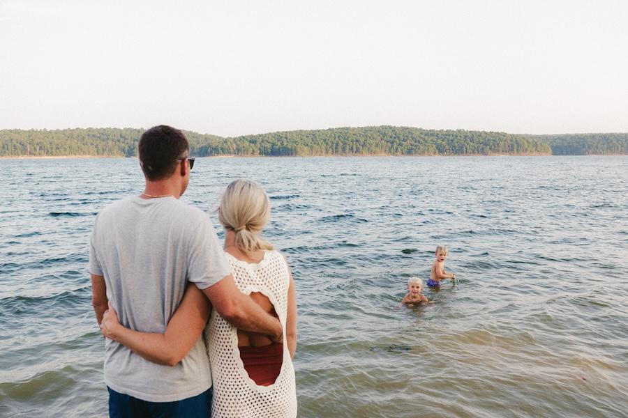 Couple embracing by a lake, with people swimming in the background.