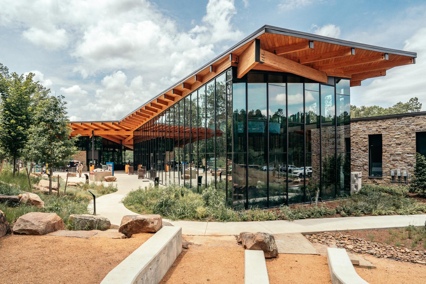 Modern building with glass walls and wooden roof in a landscaped setting.