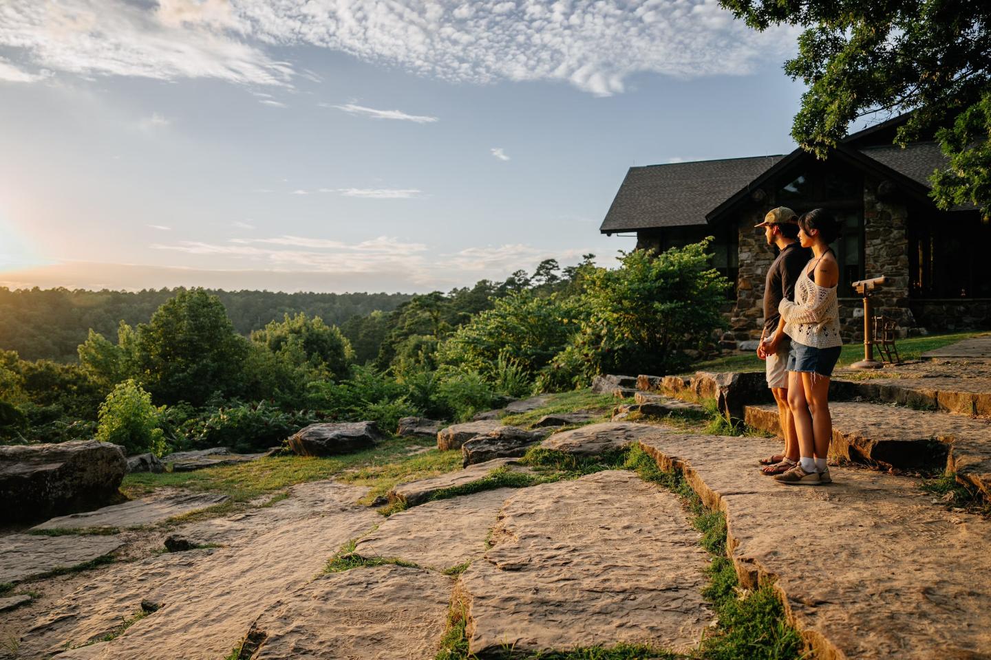 Two people stand on a rocky hill at sunset, overlooking a forest near a cabin.
