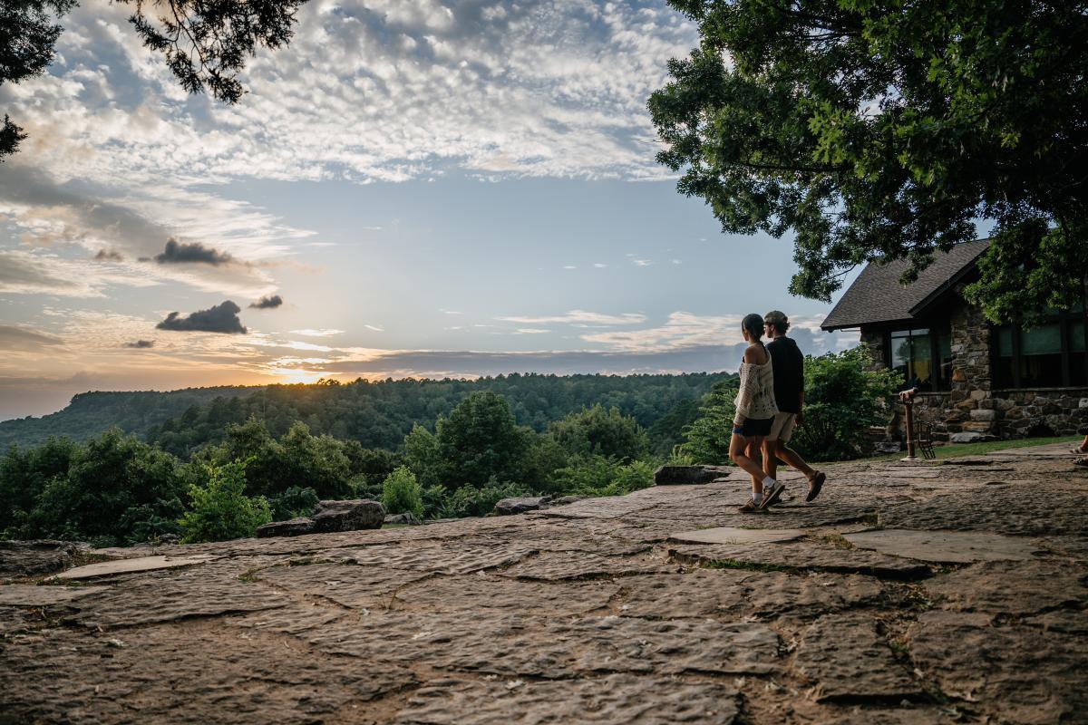 Couple walking on a rocky path towards sunset, trees and a cabin nearby.