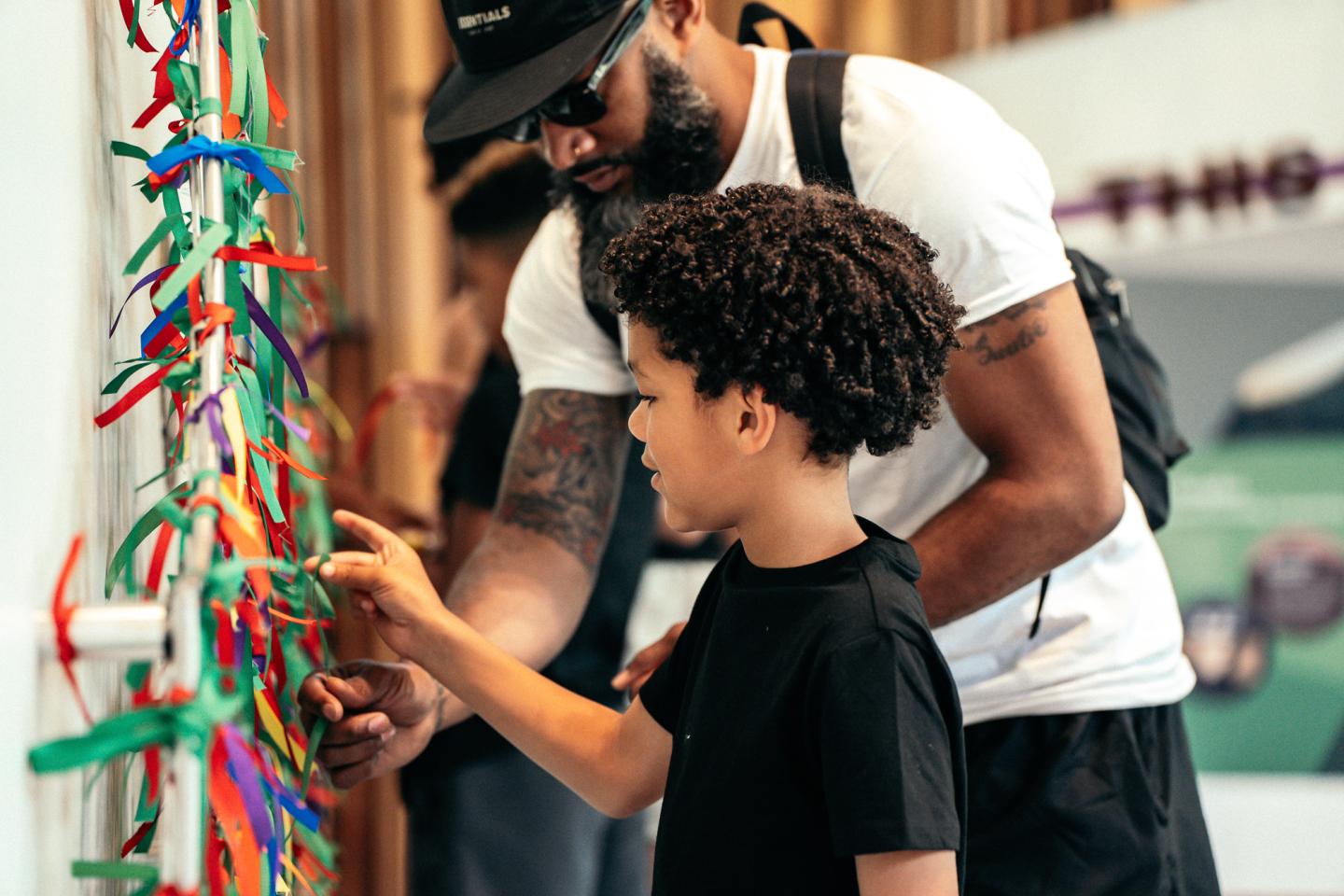 Father and son engaging with colorful ribbons on a wall exhibit.