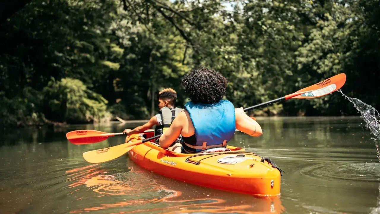 Two people kayaking on a calm river surrounded by trees.