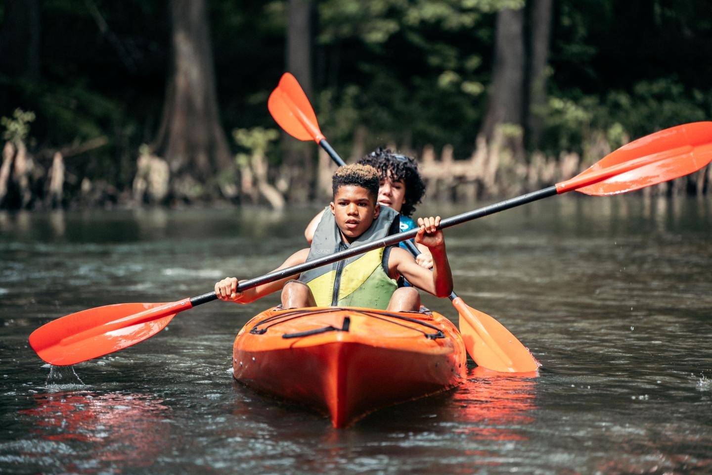 Two people kayaking on a river, paddling under a tree canopy.