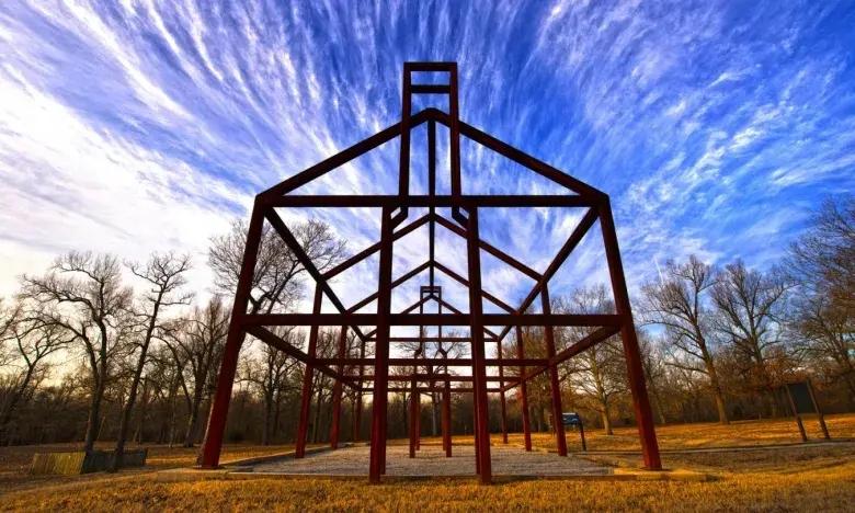 Metal house frame under a vibrant, cloud-streaked blue sky in a leafless forest.