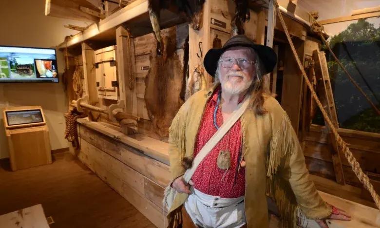 Man in pioneer attire smiles in a historical exhibit room with wooden artifacts.