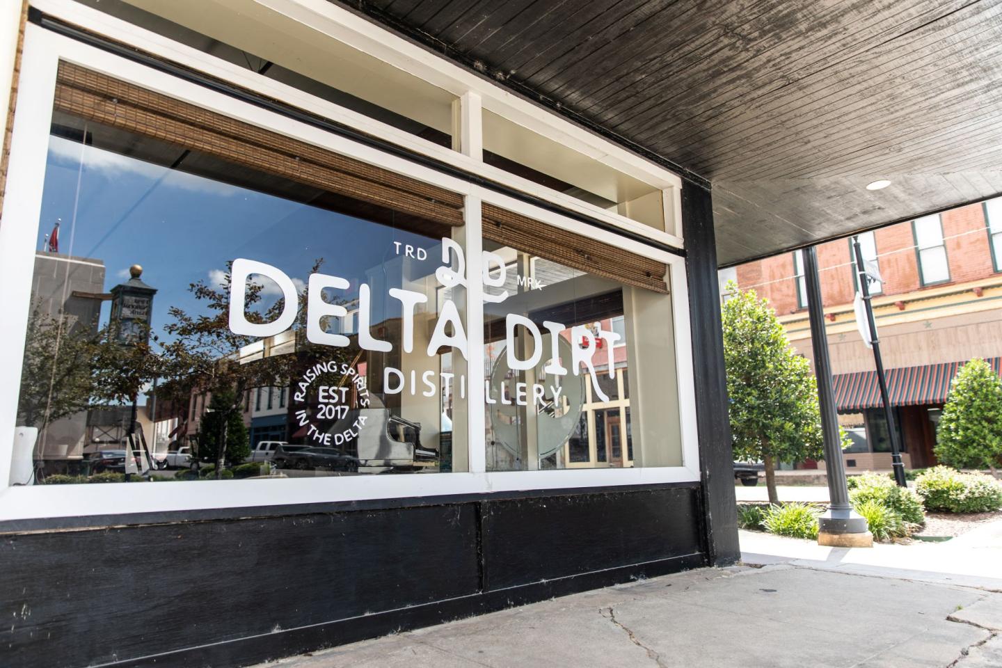 Storefront window with "Delta Dirt Distillery" logo, reflecting street scene.