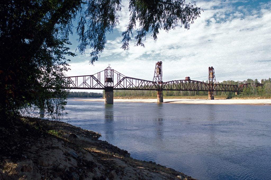 Bridge over a calm river with trees in the foreground and a cloudy sky.
