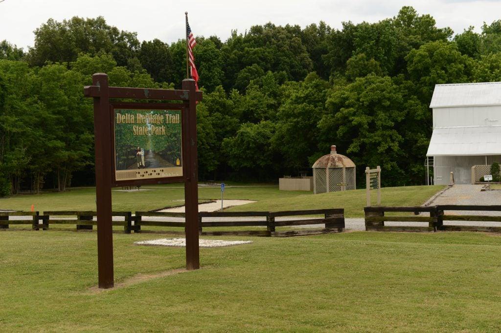 Grass park with a wooden sign, trees, and a white building in the background.