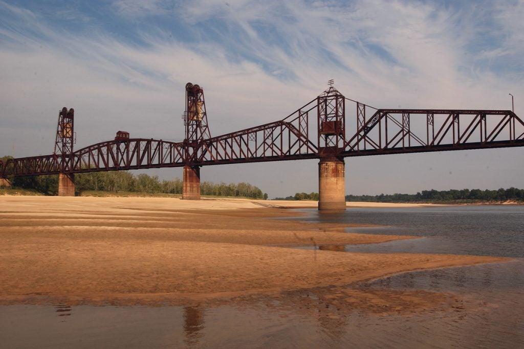 Steel bridge over sandy riverbed with cloudy sky.