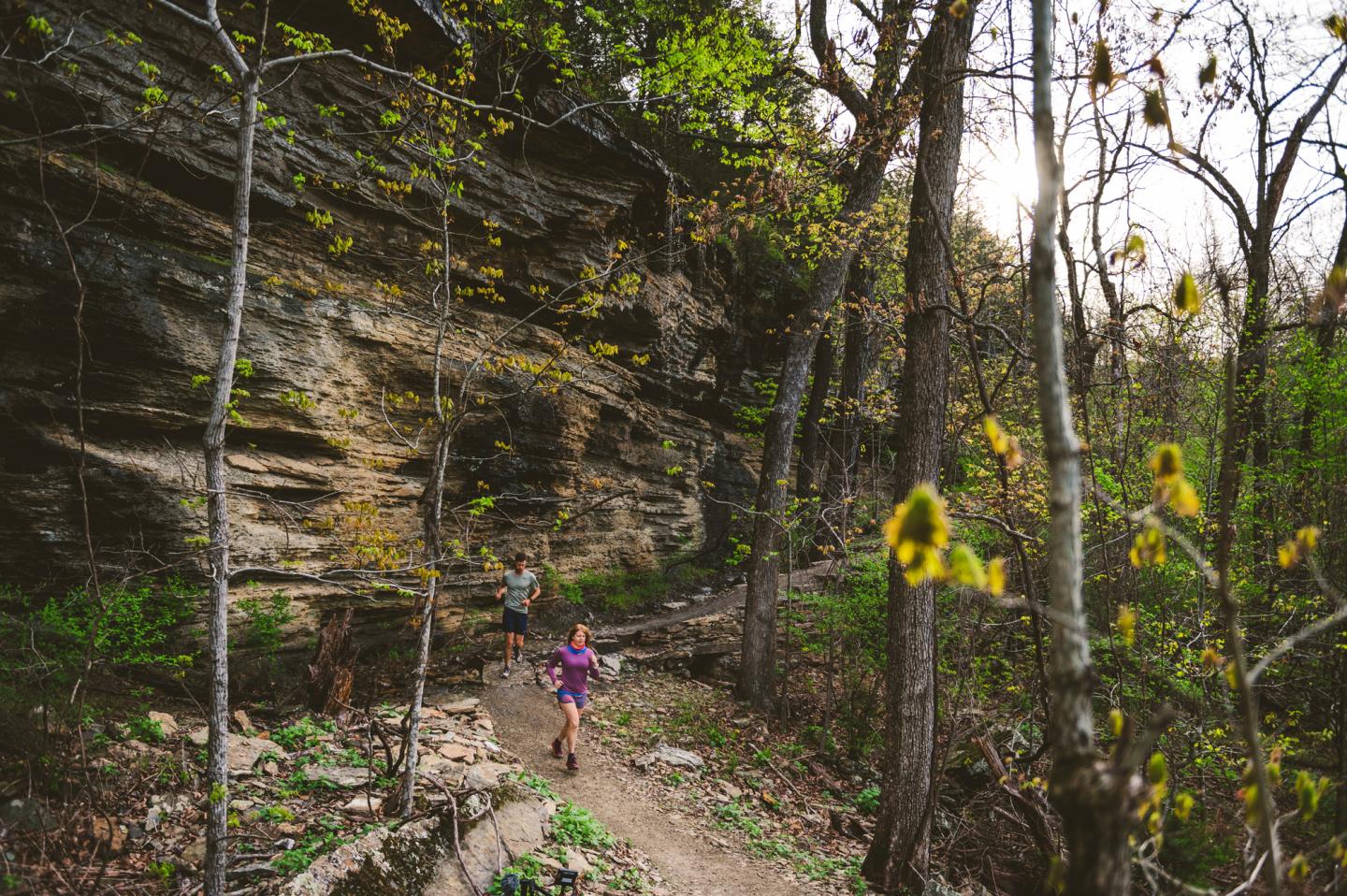 Trail runners on a forest path near a rocky cliff, sunlight filtering through trees.