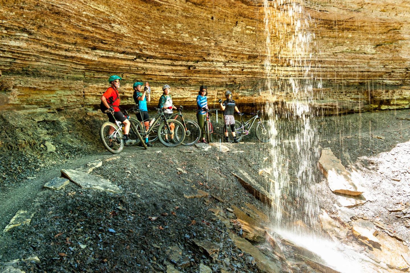 Cyclists pause by a rock cliff and waterfall on a trail.