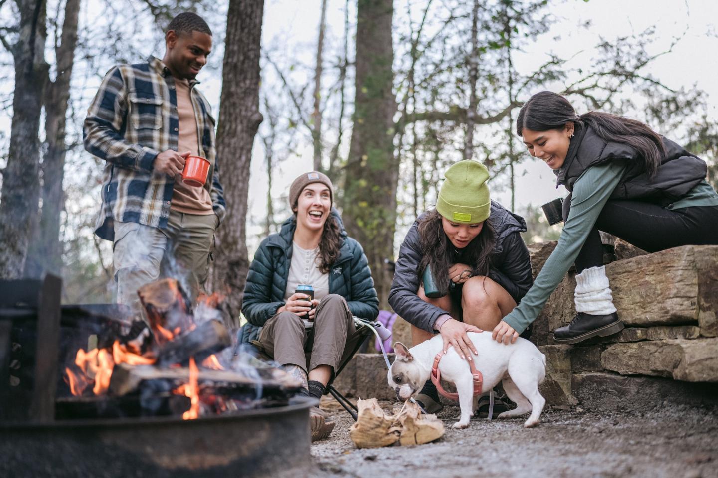 Group of friends smiling around a campfire, one petting a dog in a forest setting.