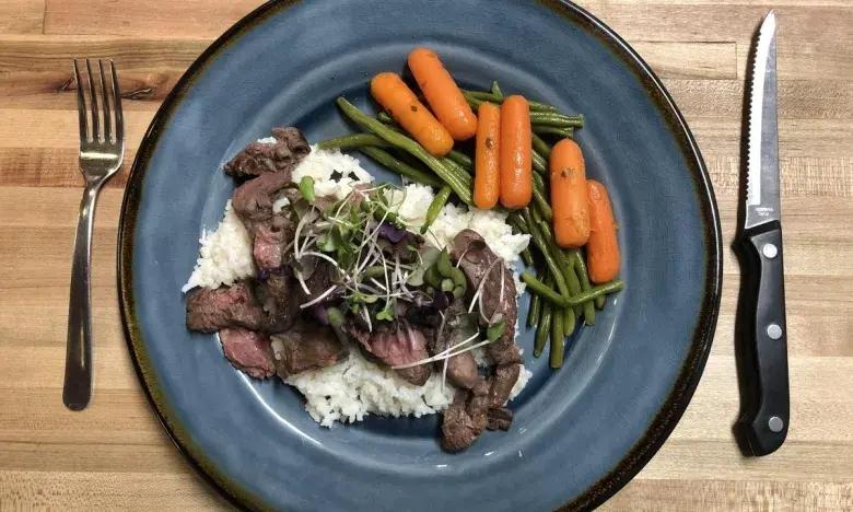 Steak with rice, garnished greens, carrots, and beans on a blue plate. Fork and knife nearby.