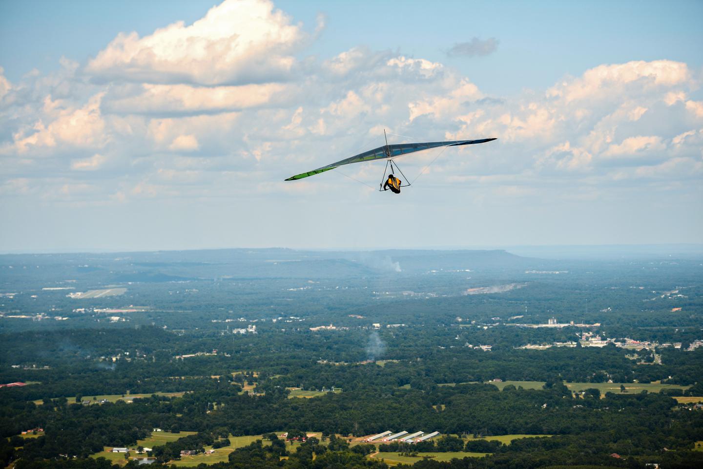 Hang glider soaring over a lush landscape with scattered clouds.