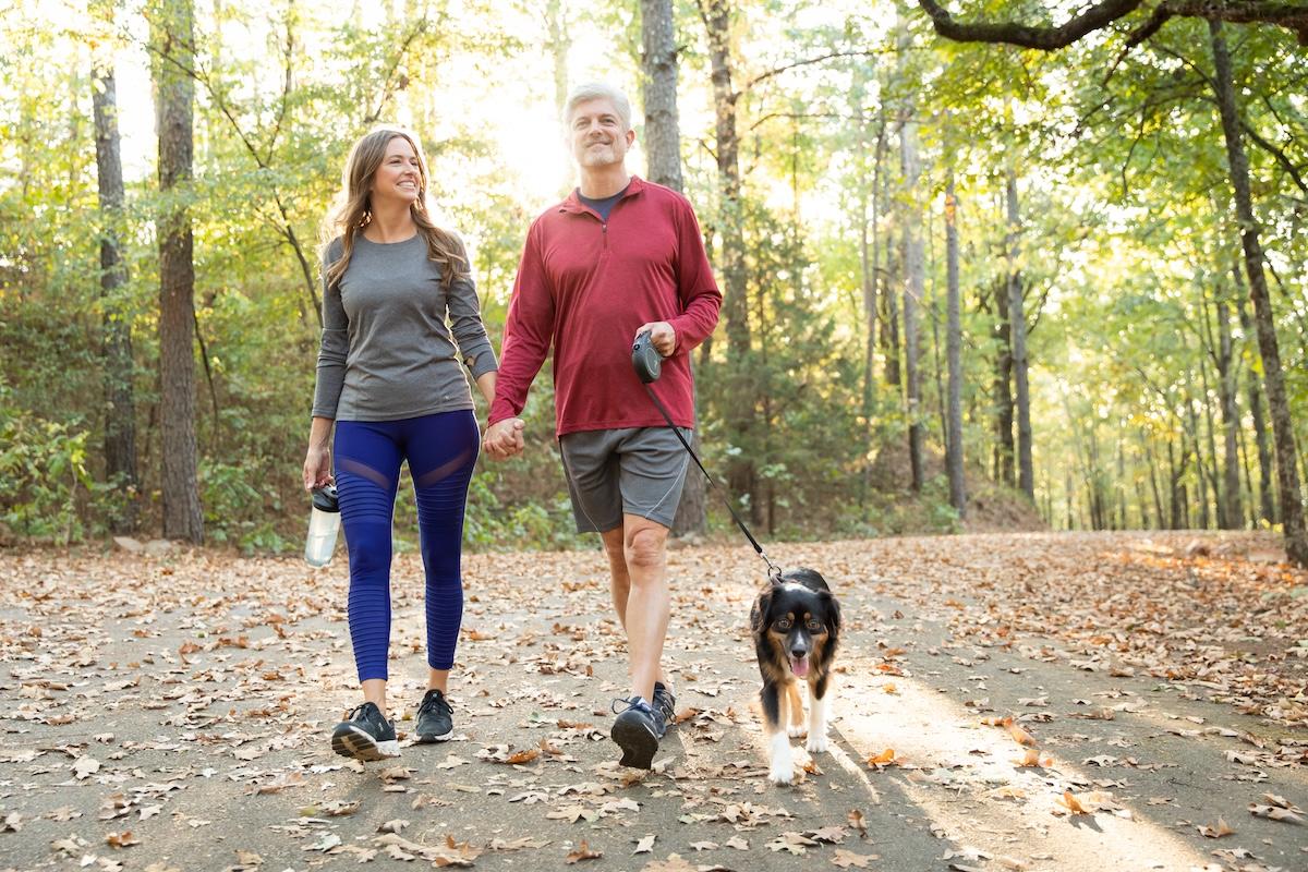 Couple walking a dog on a leaf-covered path in a sunlit forest.