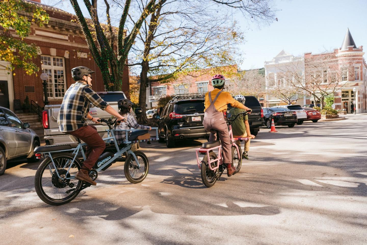 Cyclists ride through a sunny, tree-lined street with parked cars.