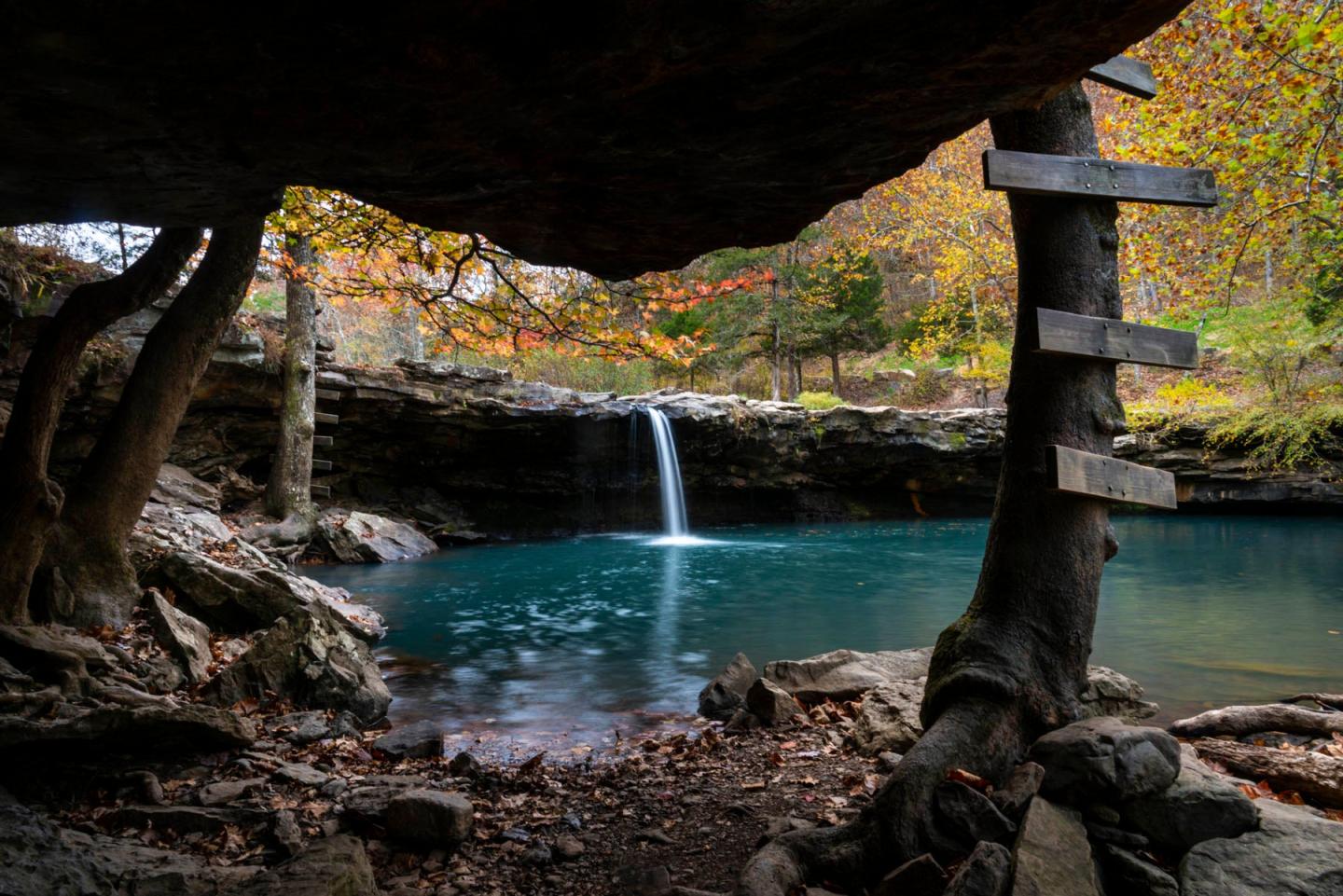 Small waterfall flowing into a turquoise pond, surrounded by autumn trees and rocks.