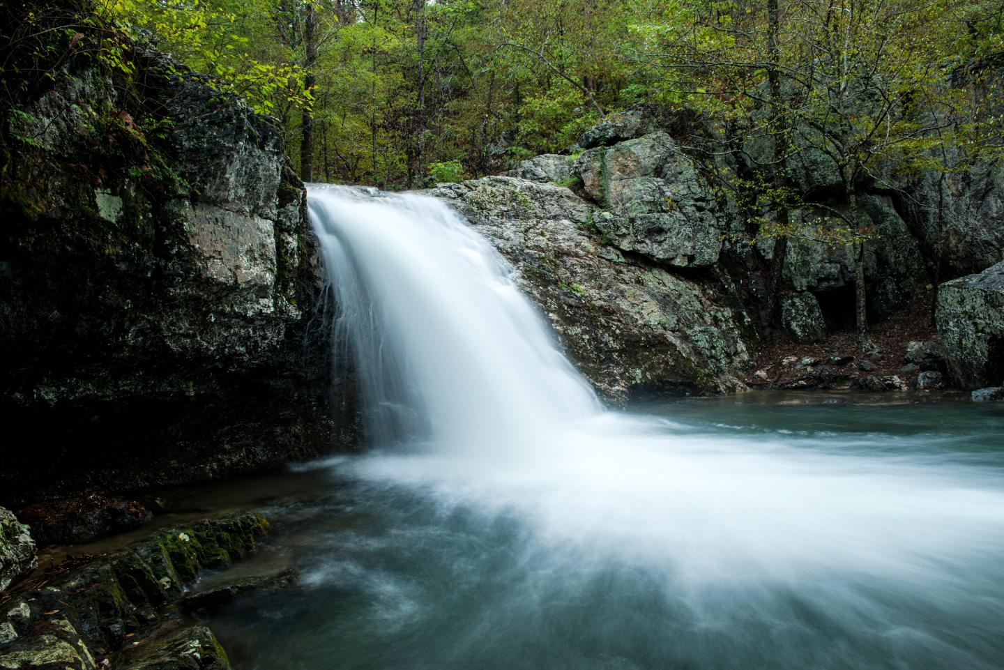 Waterfall flowing over rocks into a calm pool, surrounded by lush green forest.