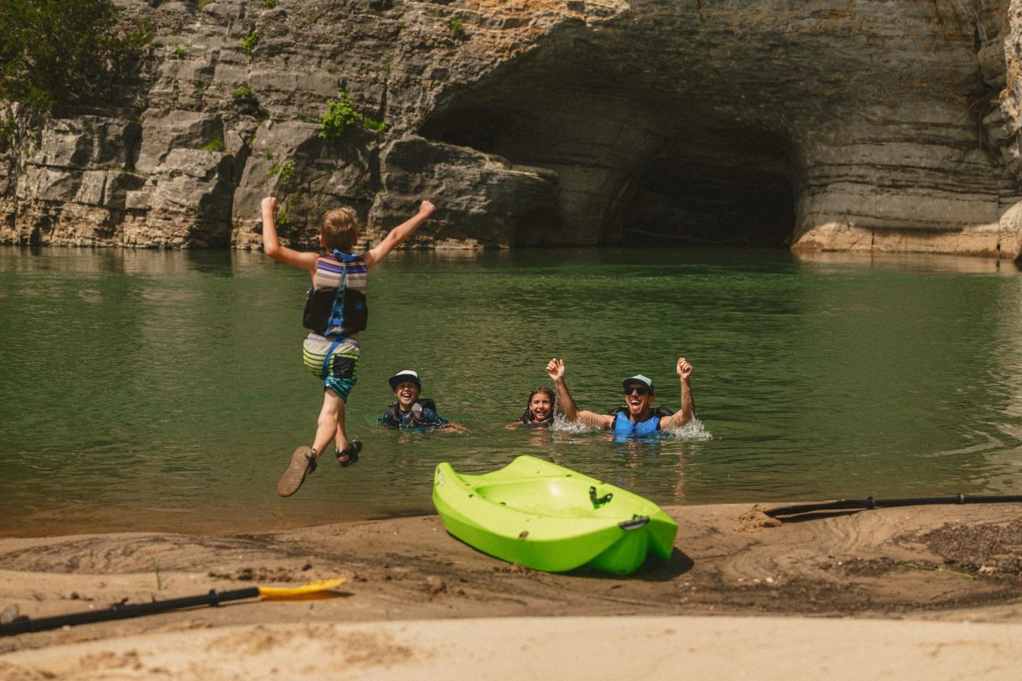 A child jumps excitedly into a green river near a cave, while others watch nearby.