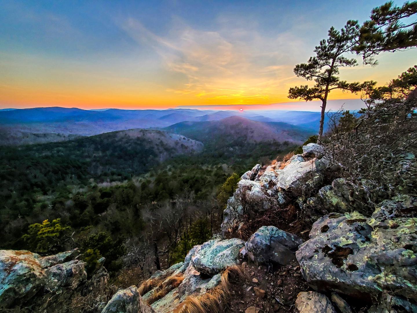 Sunset over a mountain landscape with a rocky foreground and sparse trees.