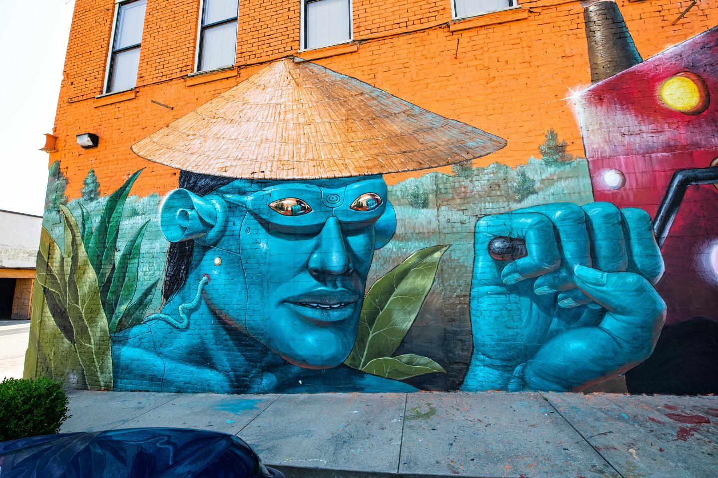 Blue-skinned figure mural on orange brick wall, wearing a straw hat and sunglasses.