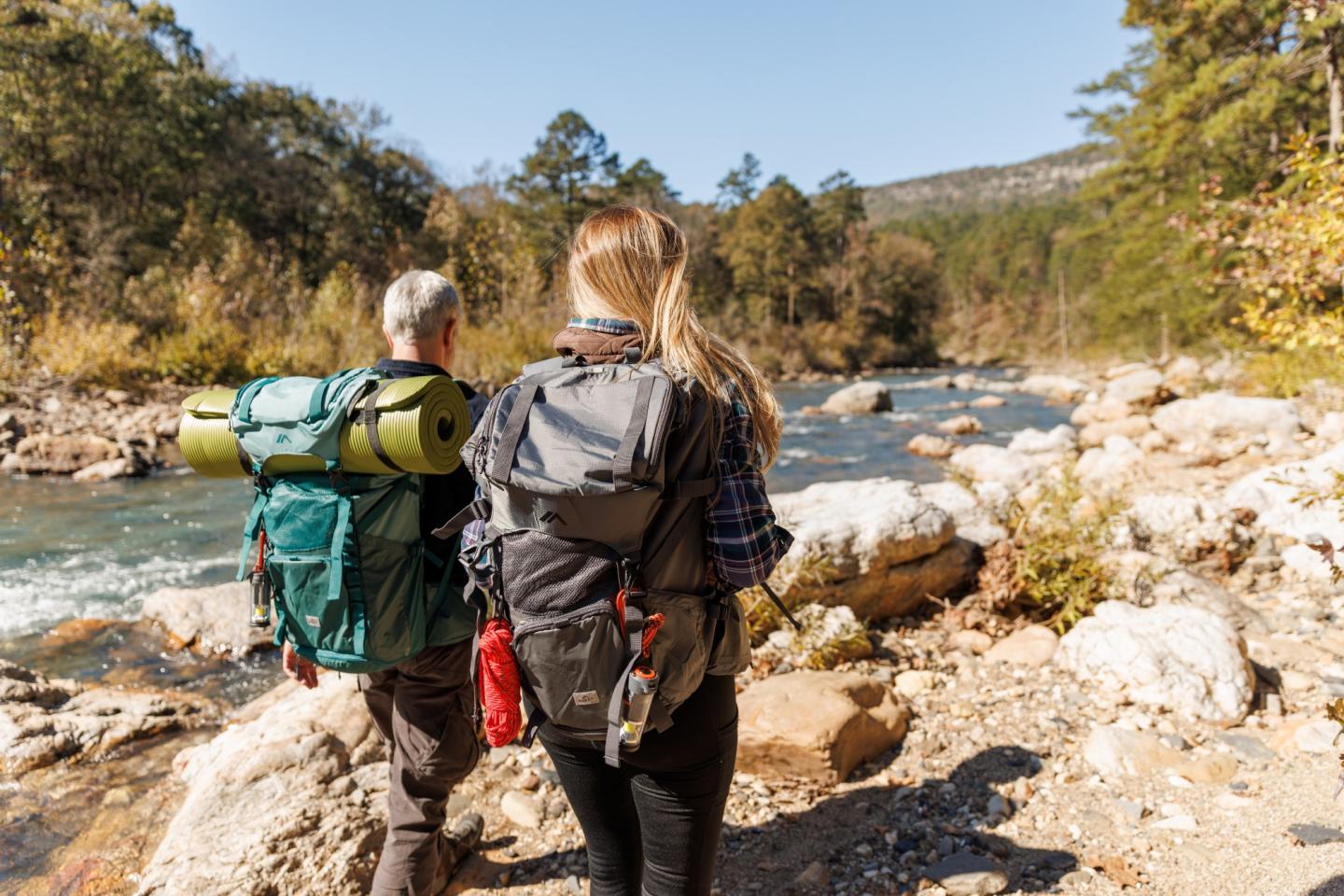 Two hikers with backpacks walking beside a scenic river in a forest.