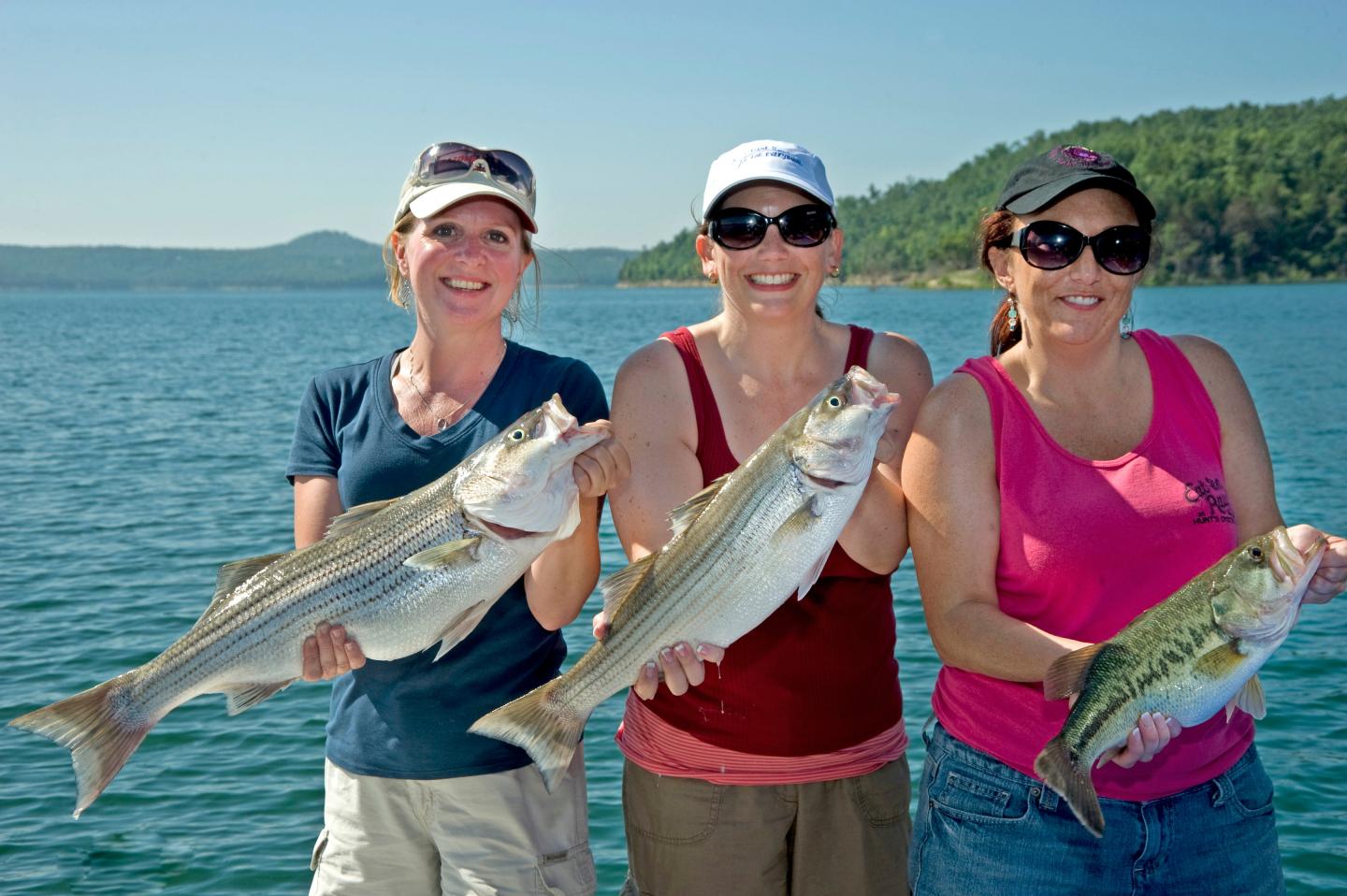 Three women smiling, holding fish on a boat with a scenic lake and trees in the background.