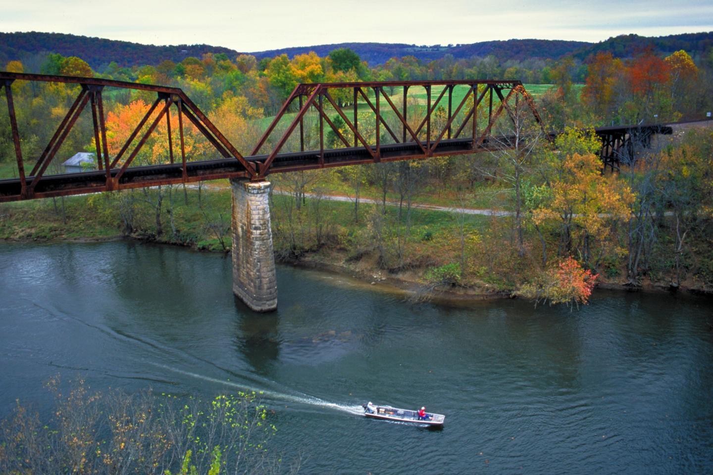 A boat on a river beneath a rusty bridge, surrounded by autumn trees.