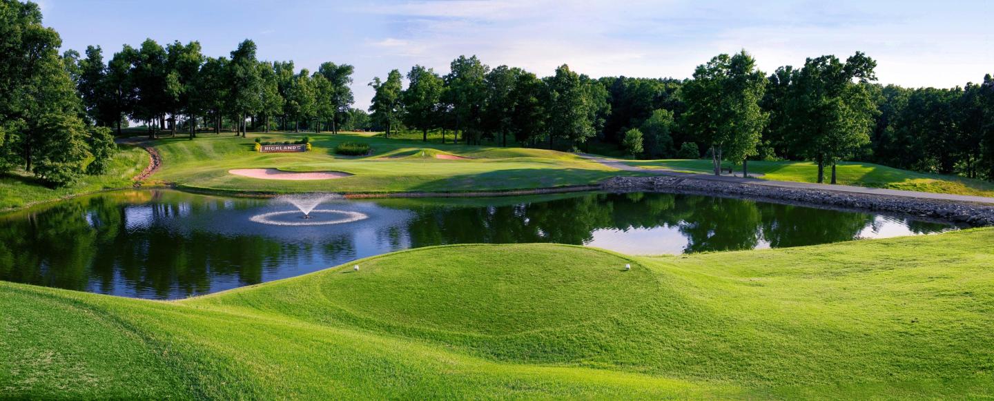 Golf course with pond, fountain, and surrounding trees under a blue sky.