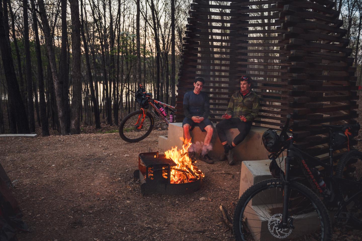 Two people sit by a campfire in a wooded area at dusk with bicycles nearby.