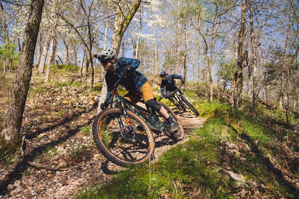 Two cyclists riding on a forest trail under bright sunlight.