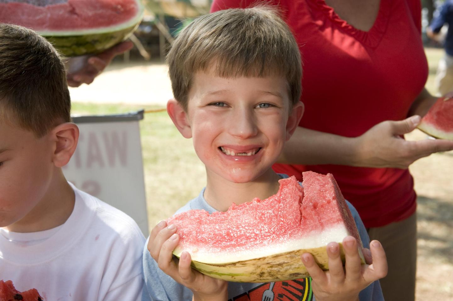 Child smiling, holding a large slice of watermelon outdoors.