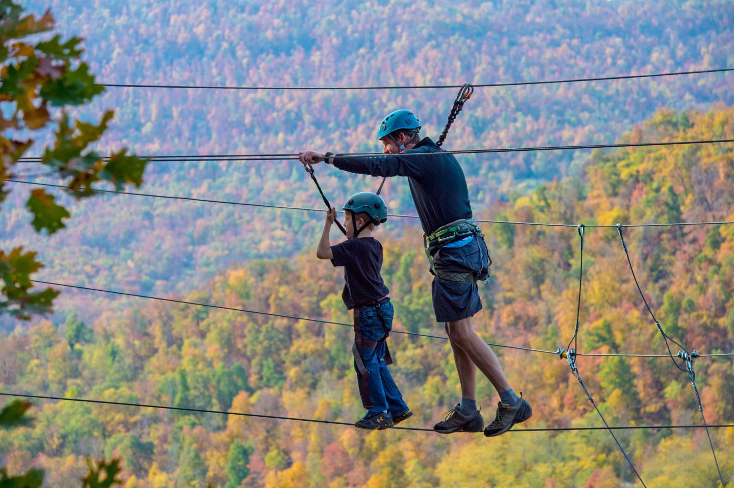 Two people on a high ropes course, wearing helmets, surrounded by autumn trees.