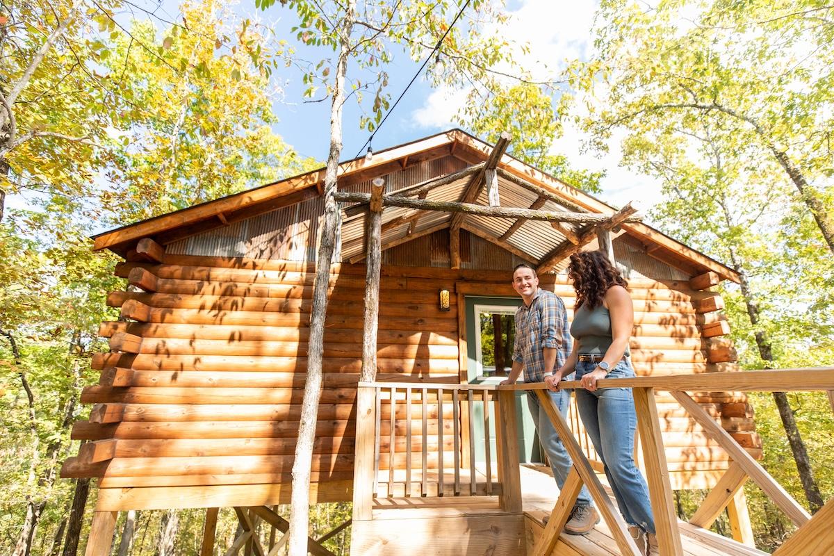 Two people smiling on a wooden cabin porch surrounded by trees.