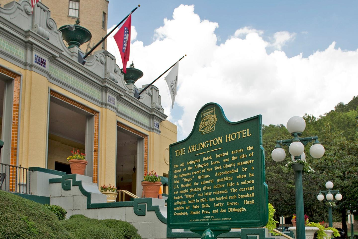 Historic hotel entrance with flags and a green informational sign, under a partly cloudy sky.